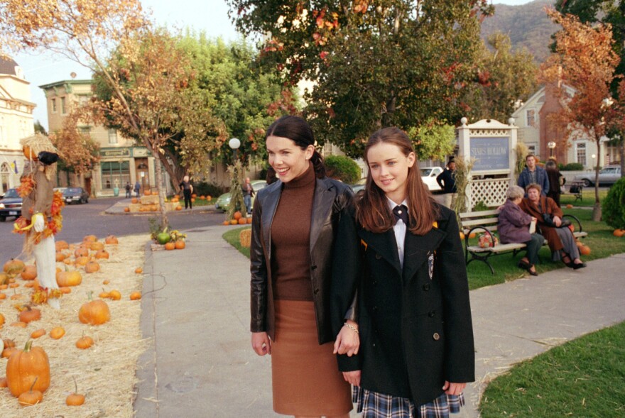 Two women stand in the center of a town square, with their arms linked. One is younger, wearing a private school uniform, the other is in a brown turtleneck, skirt and leather jacket. To their right is a scarecrow and pumpkins on hay. Behind them are trees, people on a bench, and a sign that reads "Stars Hollow."