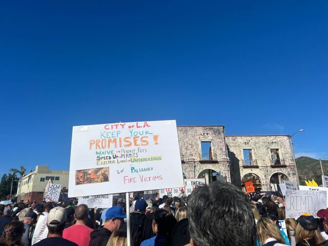 Protestors carry signs near the shell of a building in an area burned by the Palisades Fire.