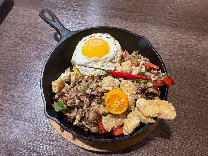 A cast iron skillet filled with sizzling sisig — a chopped pork dish with vegetables and chicharron — topped with a sunny-side-up egg, a halved calamansi and a whole red chili pepper.