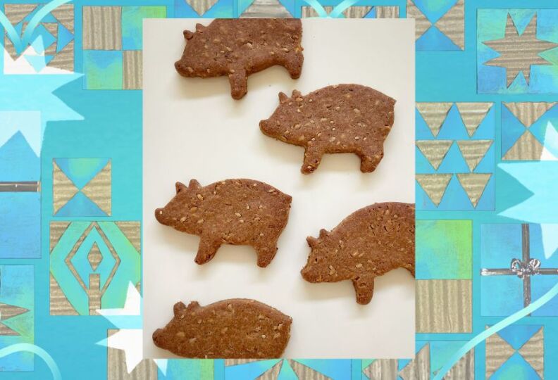 A photograph of organic dog treats that are shaped like little pigs. The treats have been baked until crispy, and are dark brown. The image is set against a decorative teal background accented with stars, gift boxes and strands of ribbon. 