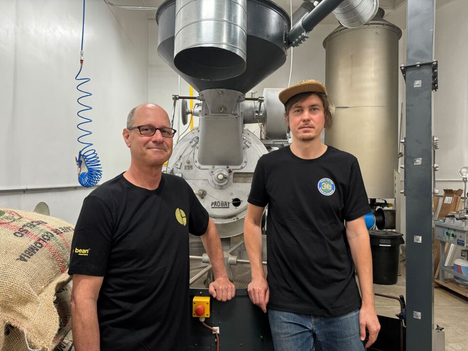 Two men, both with a light skin tone stand next to each other with a coffee roasting machine, surrounded by burlap sacks and the hum of stainless steel and ambition. One wears a well-worn ball cap, the other a quiet grin—each in black tees.