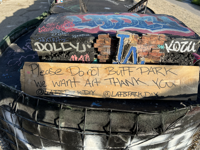 A sign on top of the front sign at the LA River Fingerboard Skatepark that says "Please Do noT BuFF PARK We WANT ART THANK YOU". Graffiti can be seen in the background.