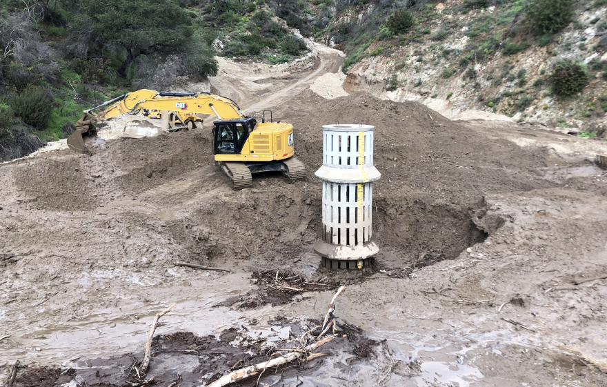 A yellow excavator moves mud away from a concrete vertical drain.