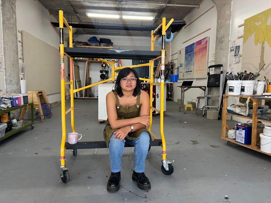 A brown skinned woman sits on a yellow dolly in the middle of a warehouse.