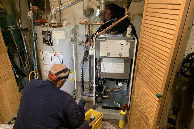 A man with light skin tone wearing headgear is crouched in front of a gas boiler.