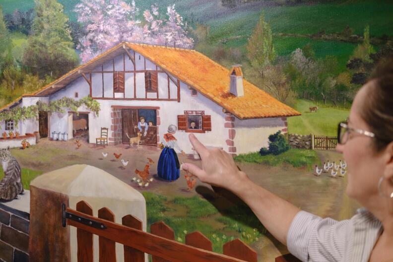 a woman points to a painting of a rustic farmhouse with a thatched roof and several chickens outside