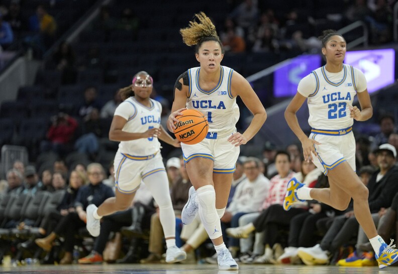Three UCLA women's basketball players run toward the camera in white, blue and yellow uniforms during a game. The player at the center oft he frame is dribbling the basketball as she goes.