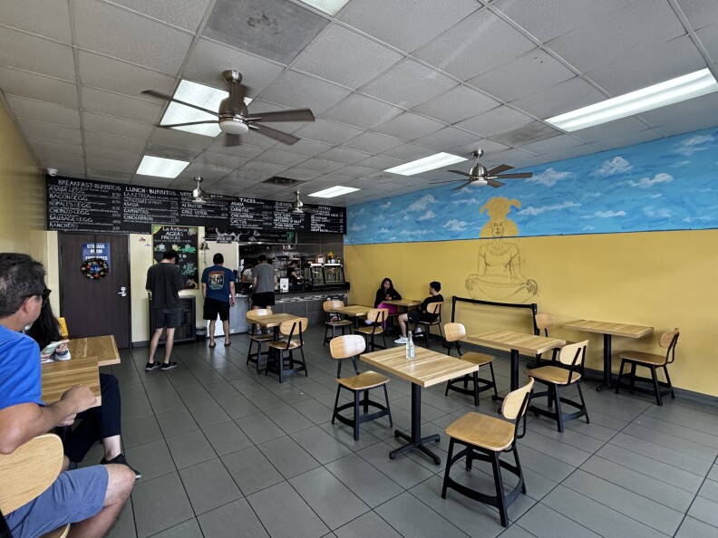 A small cafe with wooden tables and chairs spaced throughout a clean, tiled floor. A wall art mural is in progress. One wall is painted bright yellow, with a blue sky mural across the top, featuring an unfinished sketch of a seated Aztec in traditional attire. 