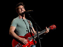 Country singer Jake Owen, a 20- or 30-something white man, smiles while standing onstage in front of a microphone. He holds a bright red guitar.