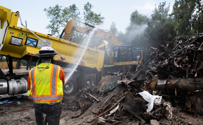 A person sprays water on piles of metal with a truck and a big machine in the background. 