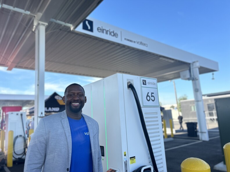 A Black man smiles wearing a gray sports jacket and blue t-shirt underneath. He stands next to a white rectangular electric vehicle charger. In the background is a large shade structure with the words "einride powered by voltera"