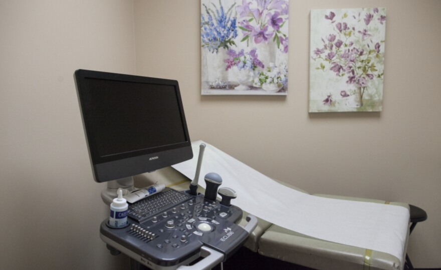 A monitor and medical imaging equipment sit next to an empty patient bed in a patient room.