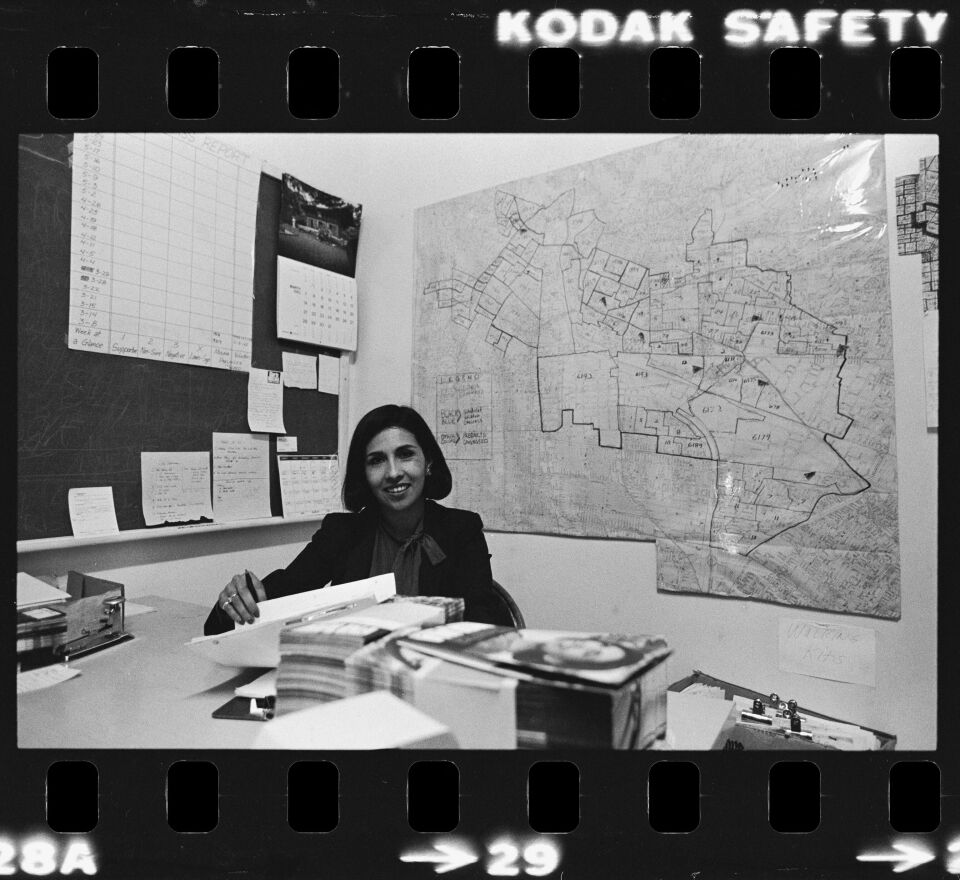 A black and white photo of a young Latina woman sitting at an office desk with stacks of paperwork. She wears a black blazer and blouse with a tied collar. Behind her there is a map with various outlines of a district. To the left of a frame there is a black board with a calendar and various sheets of paper hangin on it. On the edges of the photo you can see the film perforations and the words "Kodak"