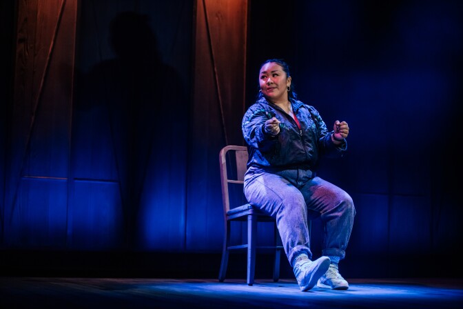 A woman with medium-light skin tone sits on a metal chair on a blue-lit stage. 