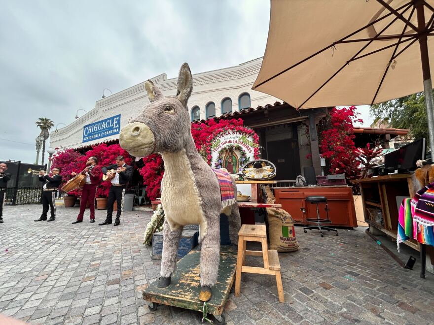 A wide shot of a large stuffed brown and tan donkey that stands outside on a platform with wheels. There are mariachis in the background and colorful plants.