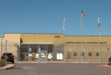 A one story, brown building behind a barbed wire fence. Three flagpoles are in front of the building. The middle flagpole is flying the American flag, the two white, unfurled flags hang on the other two poles. On the building is signage that reads, "Imperial Regional Detention Facility."