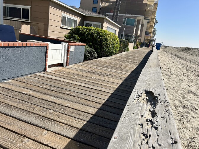 A wood walkway. Houses on the left and beach sand on the right of it.