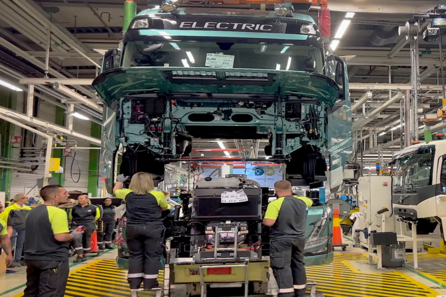 Three workers stand under a raised electric truck frame inside a factory.