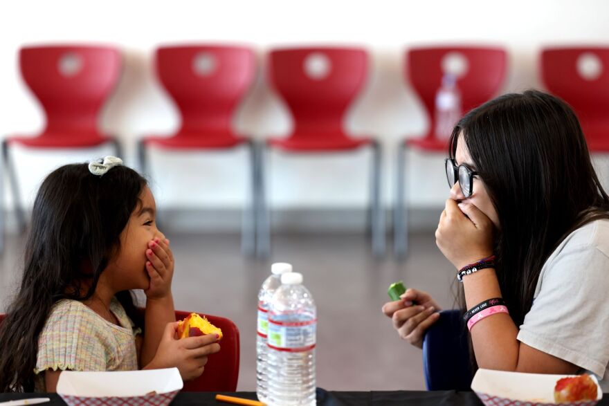 A young girl with medium skin tone and long dark hair sits on a chair behind a table. She is holding her hand to her mouth, while smiling at a woman across from her, who has long dark hair and black rim glasses and a light skin tone. The woman is also holding a hand over her mouth.