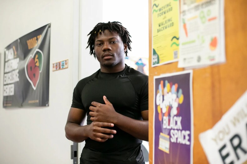 A Black male student, 18, stands in the doorway of a classroom. He is wearing a black athletic short-sleeved shirt, and he has short hair twists. He has a neutral expression, and he stands with his left hand on top of his right hand.