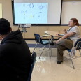 A teenage boy with medium skin tone and a black hoodie looks at a projection of math calculations. A women with medium light skin tone and glasses sits at a desk and gestures as though mid-speech. 