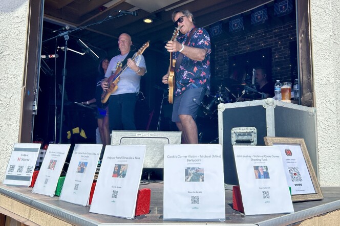 A band plays on stage behind a semi-circle of notices with QR codes to donate to victims of the Cook's Corner mass shooting. 