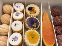 A closeup of a box of cookies including a row shaped like walnuts, another with white icing and painted flowers, and one with pressed yellow and purple flowers on top.
