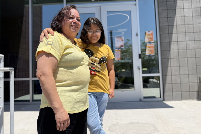 A mother wearing a yellow shirt wraps her arm around her young daughter, who is wearing a yellow Minnie Mouse shirt and glasses. They have medium skin tone and are standing in front of a building with glass doors.