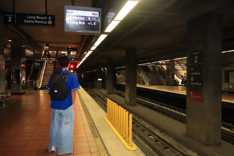 A student wearing a black backpack, blue sports jersey and blue jeans waits for a subway train on a dark subway platform.