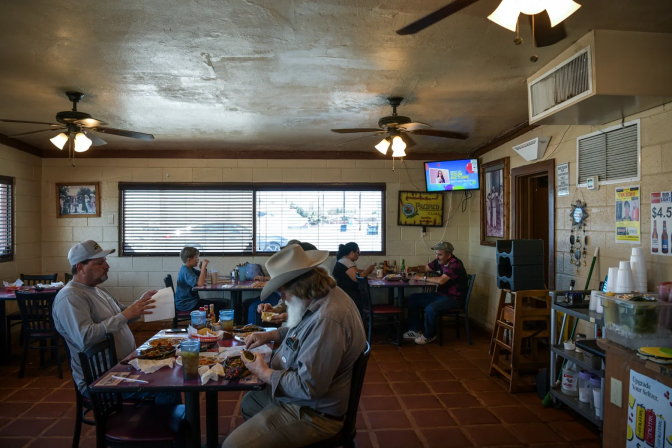 A man with a bushy white beard and wearing a Western-style hat is seated at a wooden table in a diner with another man wearing a gray long-sleeved pullover shirt and a baseball cap. A third person at the table is obscured. Other diners sit at tables in the background. A TV is mounted in a corner of the cafe.