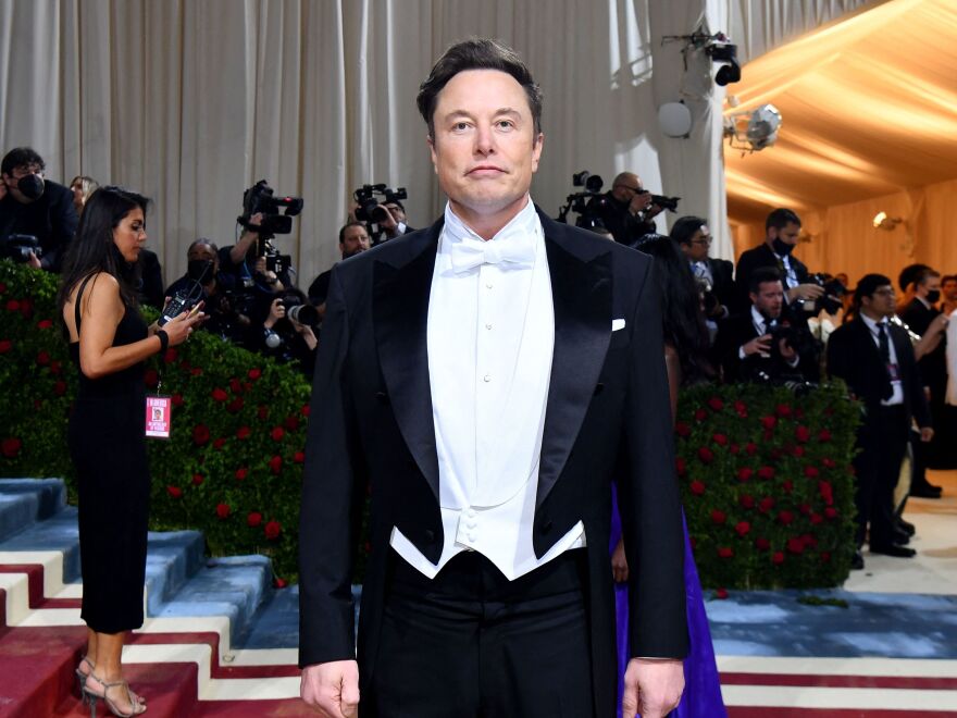 Elon Musk in a tuxedo with tails, his arms are at his side and he is not smiling.
