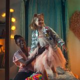 Anna Diop and Rose Decker are in a young girl's bedroom. Diop sits on the small single bed as Decker stands on top of it, Diop gentling holding her by the waist to help her balance. Both are smiling. Decker wears a pink tulle dress with a silver sequenced jacket. Diop wears a blue jeans and a red patterned top with short, flowy sleeves. The bedroom they are in is illuminated by the various string lights and lamps, giving the room a warm yellow glow. The shelves in the room are filled with books and knick-knacks. A blue curtain hangs from the wall behind Diop and Decker and a giant floral heart hangs on the wall beside it. 