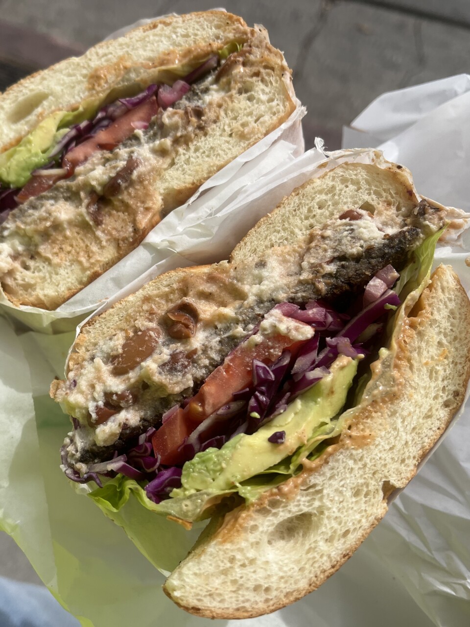 A sandwich, sliced in half to show the insides, is filled with avocado, lettuce, a large slice of red tomato, and pieces of purple cabbage. It sits on top of a thick, crumbly white substance at the bottom of a substantial roll-type bun.