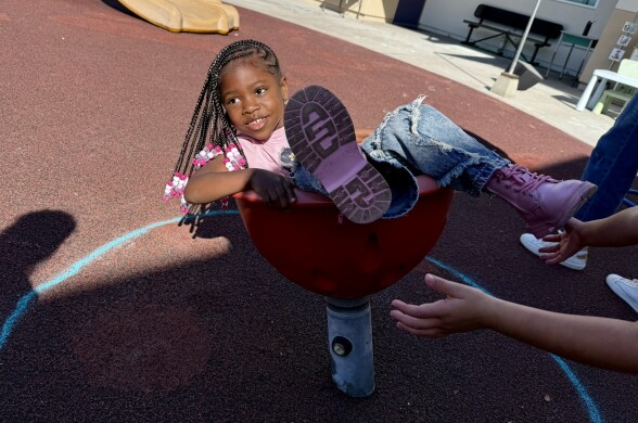  A student with dark skin tone and brown hair in braids with pink beads spins in a chair-like toy on the playground.