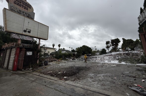 A wide view of the Hollywood Center Motel property from the sidewalk as firefighters spray water on the debris. The main home is completely demolished.