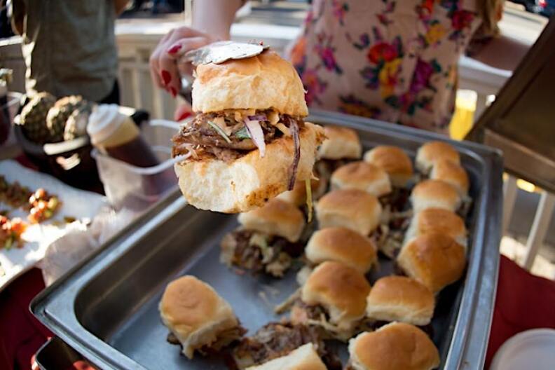 A woman wearing an apron holds uses tongs to show off one meat slider from a tray of bites.