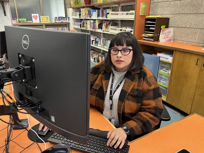 A librarian in an orange and black plaid jacket sits in front of a keyboard at an orange desk.