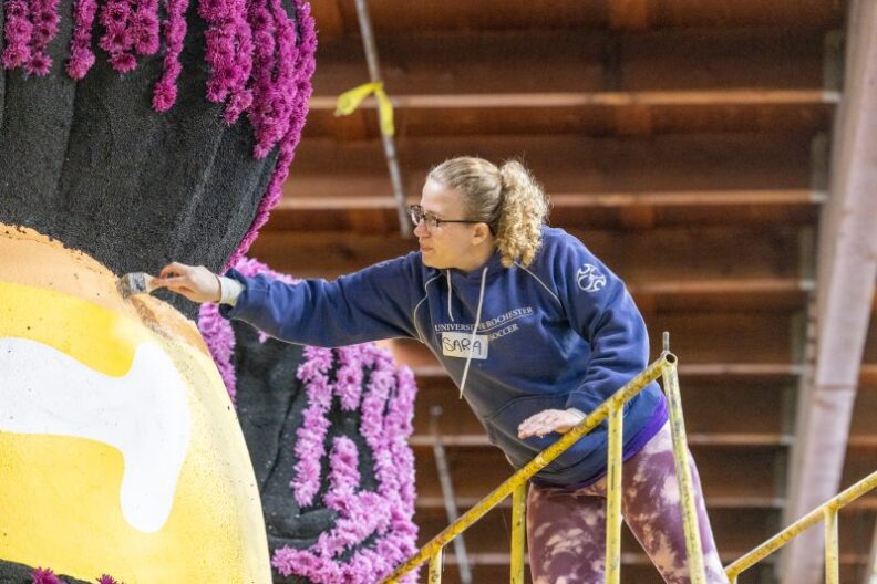 A woman wearing a blue hoodie paints a float decorated with flowers.