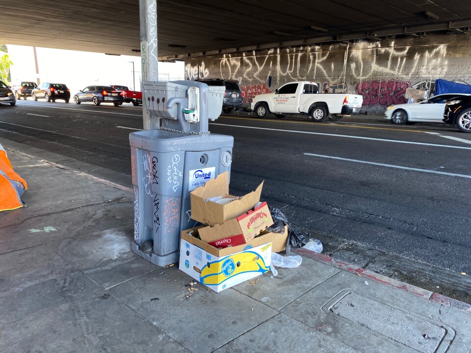 A portable handwashing station sits near an encampment under a freeway. The station is chained to a pole without a hand soap dispenser, covered in graffiti, and has boxes and trash next to it.