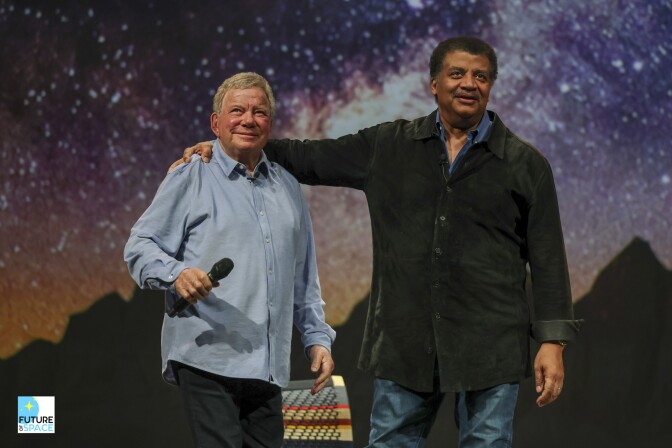 Actors William Shatner and astrophysicist Neil deGrasse Tyson smiling on stage. Shatner wears a light blue button-down and holds a microphone, while Tyson wears a dark denim shirt against a backdrop of a starry sky and mountains.