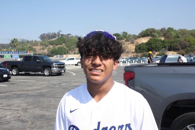 A young man of Mexican descent standing in a parking lot with a Dodgers jersey on. He has black curly hair that's partially covering his eyes with sunglasses on his head, and he is smiling. 