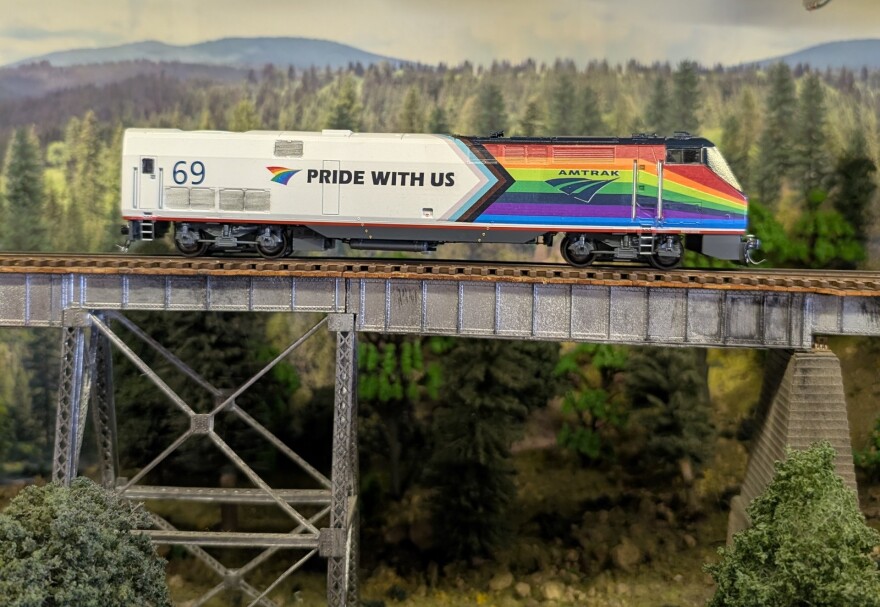 This image features a model train scene set in a detailed miniature landscape. The train is prominently displayed on a bridge and is decorated with rainbow colors, the number "69", and the phrase "PRIDE WITH US", indicating it's an Amtrak train celebrating LGBTQ+ pride. In the upper right corner, there's a small green and yellow box labeled "TrainMaster PowerMaster", which is likely part of the model train control system.