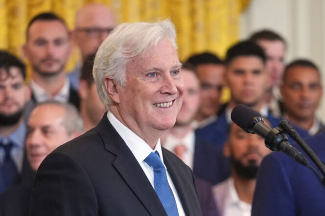 A man with white hair and a light skin tone smiles in a dark suit and blue tie, with a microphone in the foreground and a group of people in suits behind them.