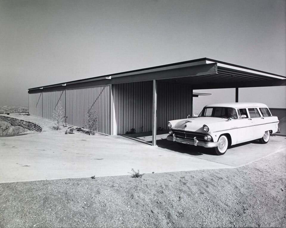 A black and white photo of a vintage car from the 1950s or 1960s parked next to a rectangular structure.
