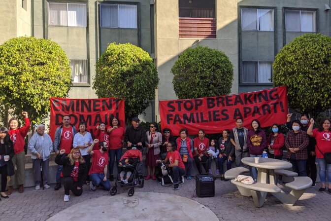 A group of tentants stand in front of a building, some with their fists in the air, with signs that say "eminent domain" and "stop breaking families apart"