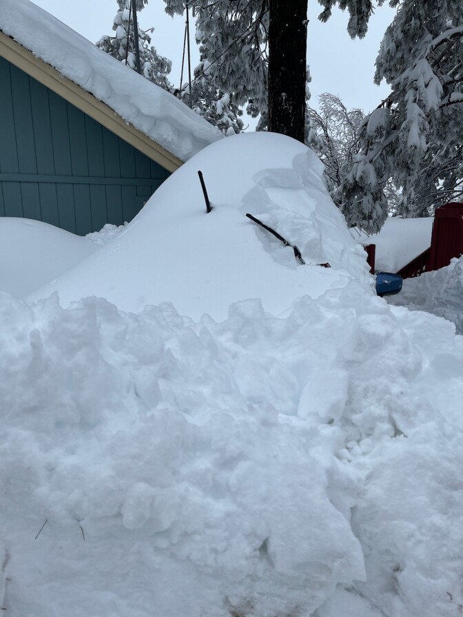 A pile of deep snow with what look like windshield wipers sticking up through it. The driver's side mirror is visible sticking out of the snow pile. There's a snow-covered angled rooftop in the background. 