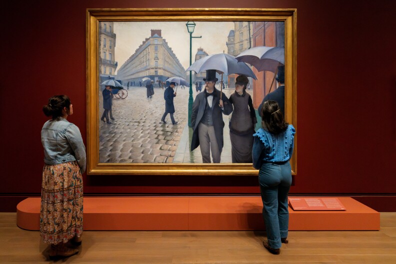 Two women stand in a gallery viewing a large Gustave Caillebotte painting of people walking in the rain with umbrellas. The painting is displayed in a gold, beveled frame on a red wall.