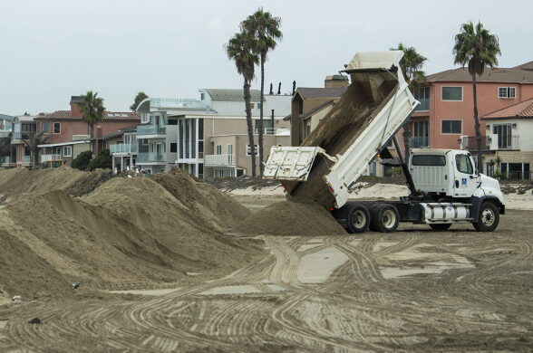 A truck dumps sand onto a beach, where a large berm rises near the water line.