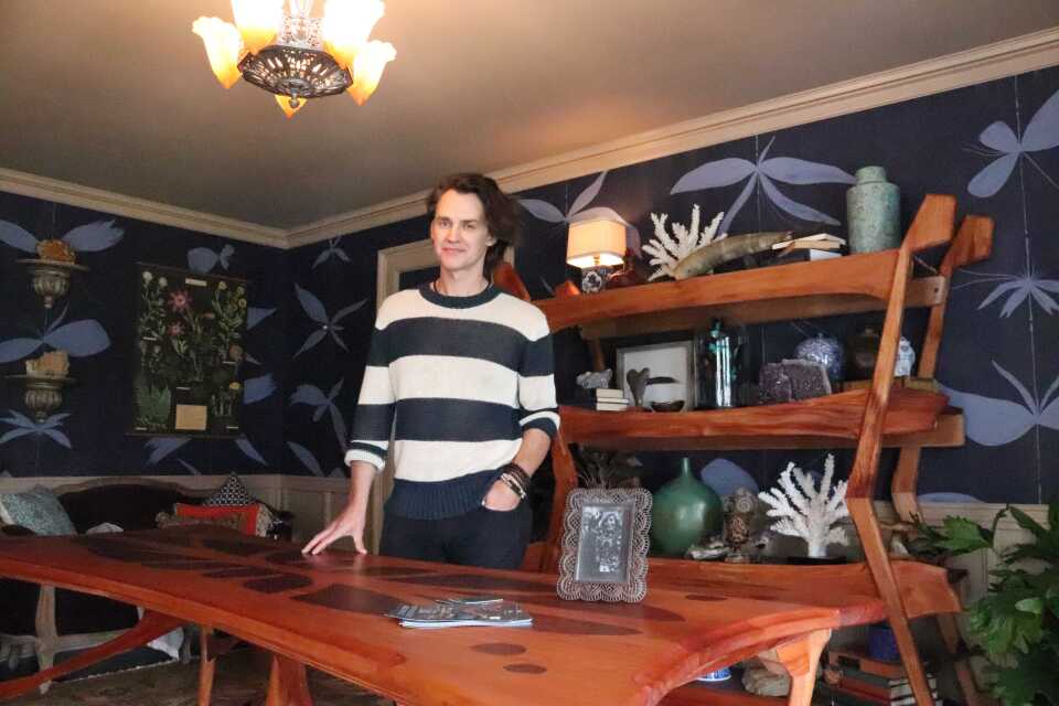 A man with fair skin who appears to be in his 30s stands behind a wood dining room table, smiling. Behind him is a large wood bookcase with art objects, a lamp and plants. 
