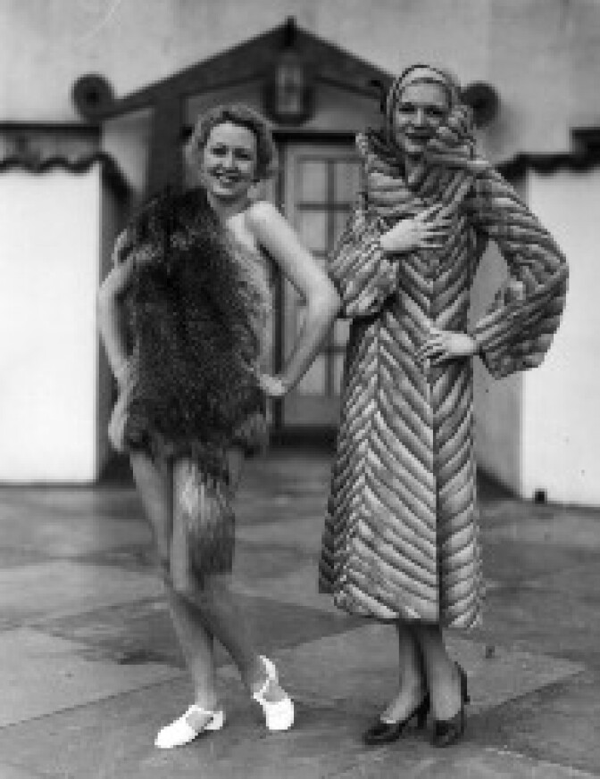 C. 1930: A fashion show including bathing suits, clothes for children, furs, and wedding dresses was held in a large Spanish-style courtyard. View 12: Young women standing next to each other model two distinctly different furs. The model on the right wears a long gray striped coat, tan hat, and black heels; the woman on the left appears to be bare underneath an animal pelt, possibly a fox fur pelt, which is draped over her right shoulder and around her hips, and wearing white shoes. 
